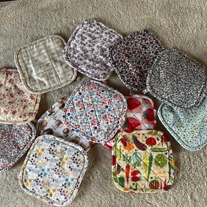 Peut inclure: Une collection de onze maniques carrées en tissu matelassé présentant divers motifs, notamment des imprimés floraux, un design à carreaux, un motif de fraises et un imprimé de légumes avec des carottes, des tomates et des aubergines.