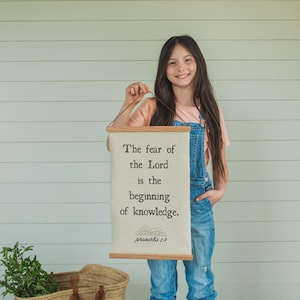 May include: A young person holds a canvas wall hanging with the text "The fear of the Lord is the beginning of knowledge." The hanging has wooden dowels and a brown cord. A woven basket with a leather handle and a bundle of greenery sits nearby.
