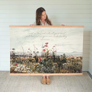 May include: A watercolor print with a quote from Proverbs 3:5, "Trust in the Lord with all your heart and lean not on your own understanding." The print features a field of wildflowers with a blue sky and a distant coastline in the background.