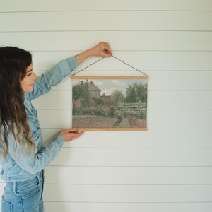 May include: A framed landscape print with a wooden hanger. The artwork depicts a garden scene with a house in the background. The print is being held against a white wall.