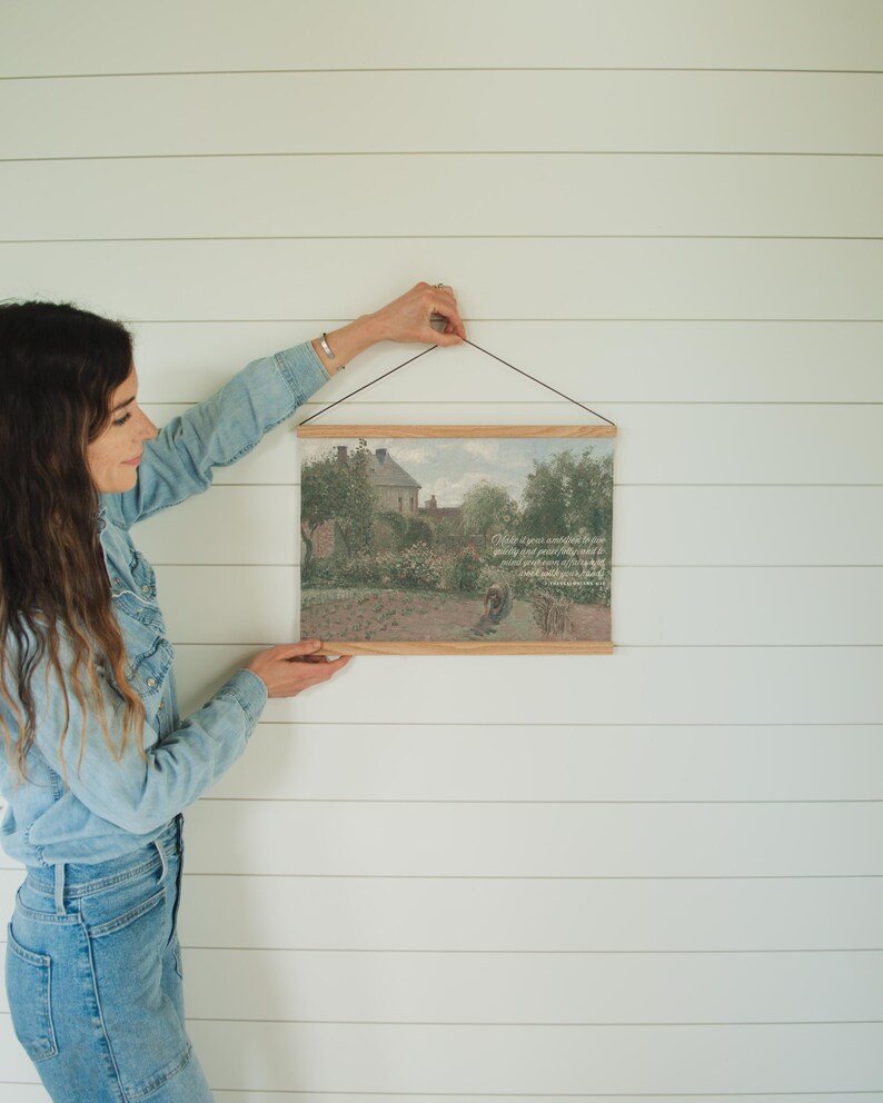May include: A framed landscape print with a wooden hanger. The artwork depicts a garden scene with a house in the background. The print is being held against a white wall.