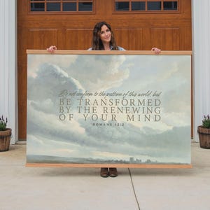 May include: A large rectangular canvas print with a wooden frame. The artwork depicts a cloudy sky with a landscape at the bottom. Text reads "Do not conform to the pattern of this world, but BE TRANSFORMED BY THE RENEWING OF YOUR MIND ROMANS 12:2."