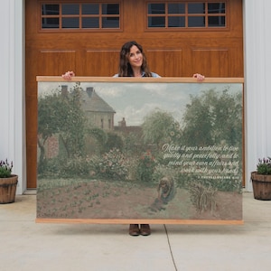 May include: A framed landscape painting with a wooden frame. The artwork depicts a garden scene with a house in the background. Text on the painting reads: "Make it your ambition to live quietly and peacefully... work with your hands."
