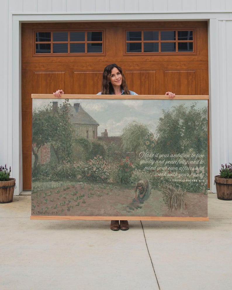 May include: A framed landscape painting with a wooden frame. The artwork depicts a garden scene with a house in the background. Text on the painting reads: "Make it your ambition to live quietly and peacefully... work with your hands."