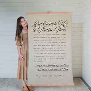 May include: A beige canvas wall hanging with wooden dowels at the top and bottom. The text "Lord Teach Me to Praise Thee" is displayed in elegant script. The quote is from St. Augustine of Hippo, Confessions, AD 400.