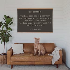 May include: A brown leather sofa with a dog sitting on it, a throw blanket, and a white pillow. Above the sofa is a framed sign with the text "THE BLESSING" and a Bible verse. A potted plant is to the left.