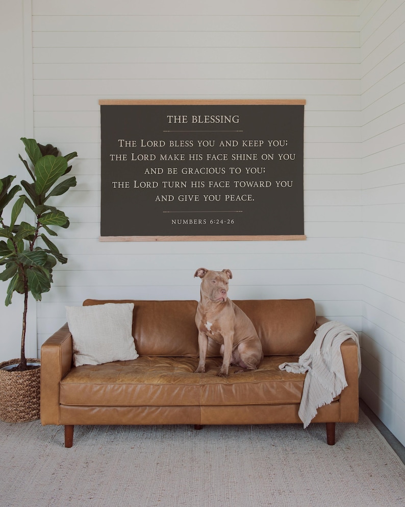 May include: A brown leather sofa with a dog sitting on it, a throw blanket, and a white pillow. Above the sofa is a framed sign with the text "THE BLESSING" and a Bible verse. A potted plant is to the left.