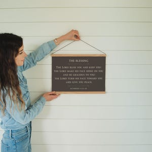 May include: A framed wall hanging with the text "THE BLESSING" and a Bible verse from Numbers 6:24-26. The text is in a dark color on a rectangular background, with wooden bars at the top and bottom. The wall hanging is suspended by a black string.