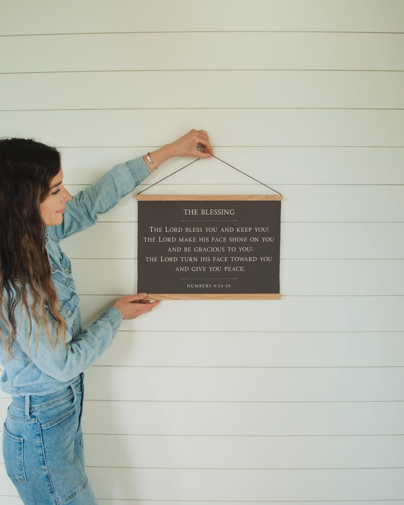 May include: A framed wall hanging with the text "THE BLESSING" and a Bible verse from Numbers 6:24-26. The text is in a dark color on a rectangular background, with wooden bars at the top and bottom. The wall hanging is suspended by a black string.