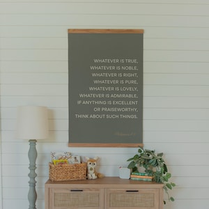 May include: A framed wall hanging with a wooden top and bottom, displaying the text "WHATEVER IS TRUE, WHATEVER IS NOBLE...THINK ABOUT SUCH THINGS." Below, a wooden dresser with a woven front, a lamp, and decorative items.