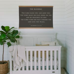 May include: A white crib with a neutral-toned blanket and pillows. Above the crib hangs a framed sign with the text "THE BLESSING" and a Bible verse. A potted plant in a woven basket sits to the left.