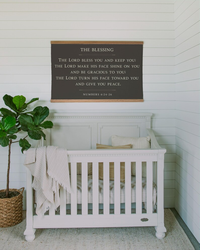 May include: A white crib with a neutral-toned blanket and pillows. Above the crib hangs a framed sign with the text "THE BLESSING" and a Bible verse. A potted plant in a woven basket sits to the left.