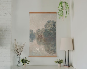 May include: A wall hanging with a landscape painting, featuring a serene water scene and trees, is displayed. The text "BE STILL and know" is printed on the artwork. A white cabinet with carved doors, a lamp, and a hanging plant complete the decor.