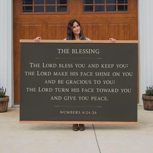 May include: A large, rectangular wooden sign with the text "THE BLESSING" and a Bible verse from Numbers 6:24-26 in white lettering. The sign has a dark gray background and a natural wood frame. The sign is held by a person.