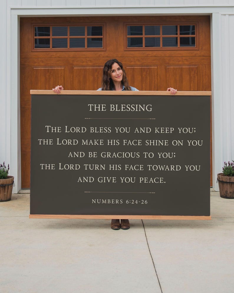 May include: A large, rectangular wooden sign with the text "THE BLESSING" and a Bible verse from Numbers 6:24-26 in white lettering. The sign has a dark gray background and a natural wood frame. The sign is held by a person.