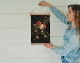 May include: A person hangs a framed floral print on a white wall. The print features a bouquet of pink and red roses on a dark background.