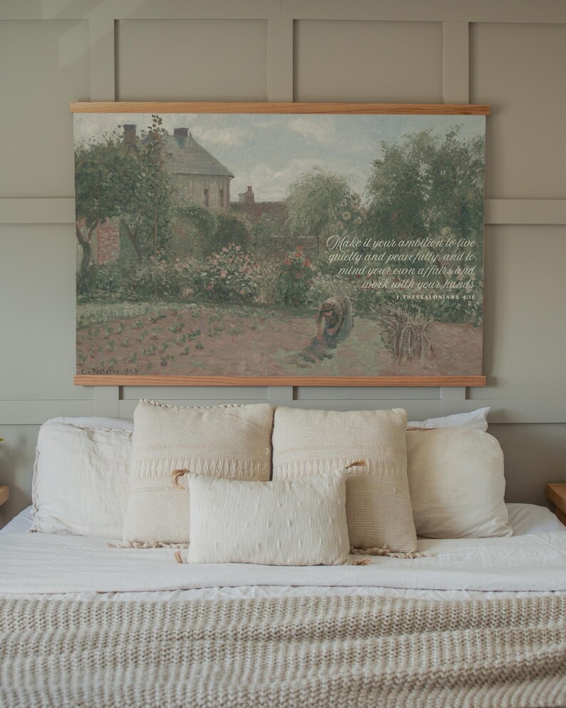 May include: A bedroom scene featuring a bed with white bedding and several cream-colored pillows. Above the bed hangs a framed landscape painting with text that reads "Make it your ambition to live quietly and peacefully, and to mind your own affairs and work with your hands."