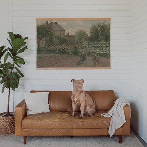May include: A tan leather sofa with wooden legs, a white pillow, and a light gray throw blanket. A large dog sits on the sofa. Above the sofa is a framed landscape print with text.