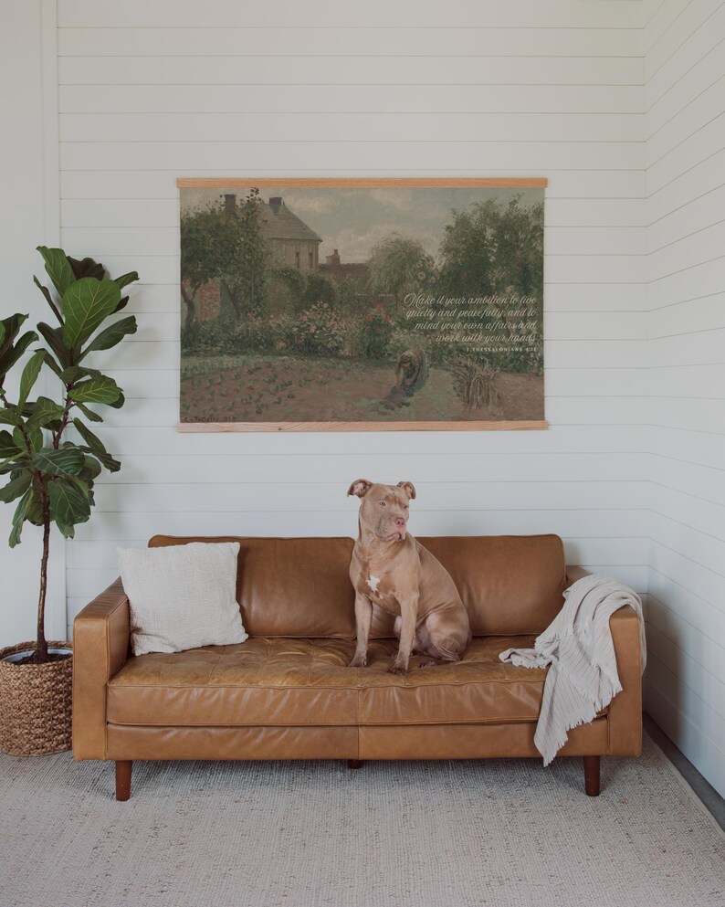 May include: A tan leather sofa with wooden legs, a white pillow, and a light gray throw blanket. A large dog sits on the sofa. Above the sofa is a framed landscape print with text.