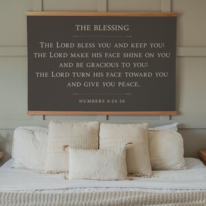 May include: A framed wall hanging with the text "THE BLESSING" and a Bible verse from Numbers 6:24-26. The dark gray sign is mounted on a wooden frame and hangs above a bed with white bedding and decorative pillows.