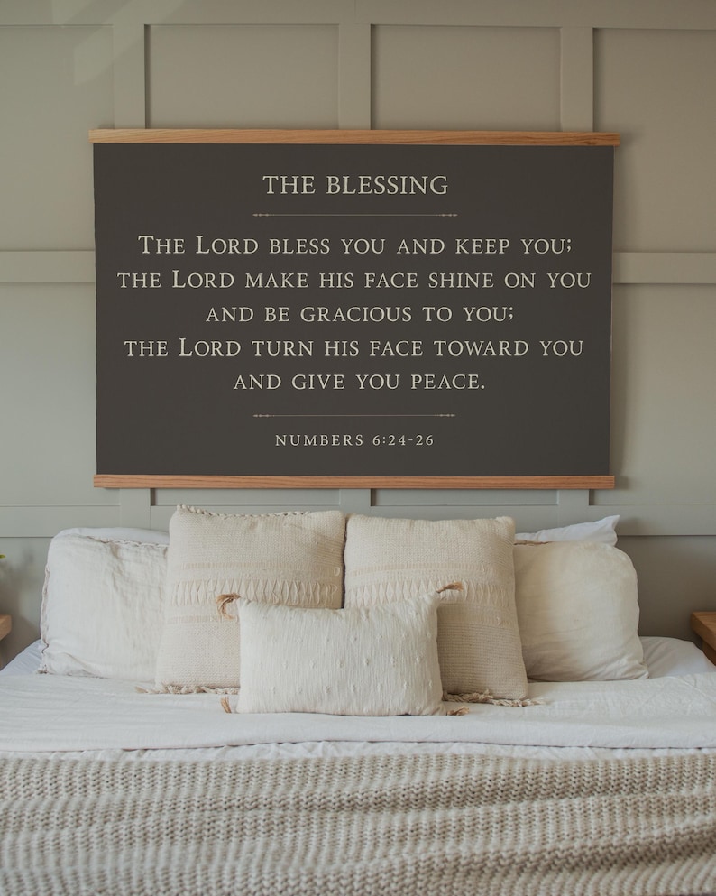 May include: A framed wall hanging with the text "THE BLESSING" and a Bible verse from Numbers 6:24-26. The dark gray sign is mounted on a wooden frame and hangs above a bed with white bedding and decorative pillows.