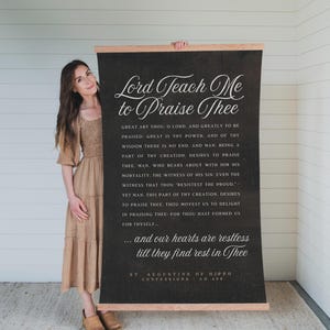May include: A large, black canvas wall hanging with the text "Lord Teach Me to Praise Thee" in elegant white script. The text is a quote from St. Augustine of Hippo. The canvas is framed with wooden rods.