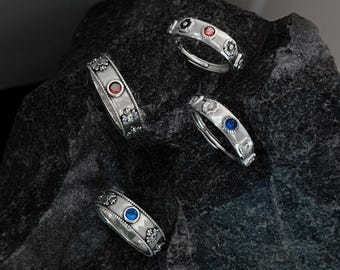 Könnte beinhalten: Fünf silberne Ringe mit floralen Designs und bunten Edelsteinen. Die Ringe haben rote, blaue und schwarze Edelsteine.