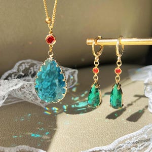 May include: A gold-toned necklace with a large, teardrop-shaped, teal-colored pendant and a small red accent stone. Matching gold earrings with green teardrop-shaped stones and red accents are also displayed. The jewelry is set against a soft, neutral background.