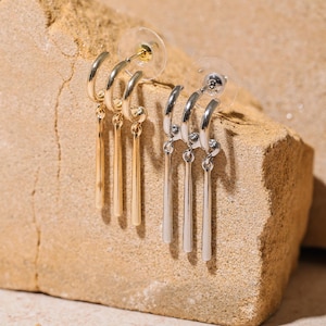 May include: A pair of gold hoop earrings with three vertical bar pendants and a pair of silver hoop earrings with three vertical bar pendants. The earrings are displayed on a textured, light brown surface.