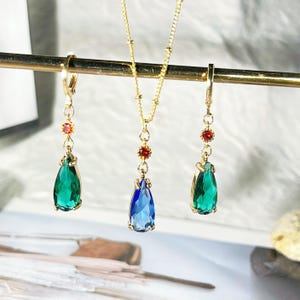 May include: A gold necklace and earrings set with teardrop-shaped green and blue gemstones. The necklace has a delicate chain with small beads and the earrings have a simple hook closure.
