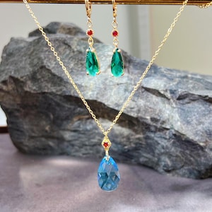 May include: A gold-coloured jewellery set comprising a necklace and earrings. The necklace features a teardrop-shaped blue pendant with a red accent. The earrings have green teardrop-shaped gems with red accents. The jewellery is set against a grey stone background.