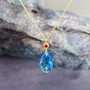Op de afbeelding: Een goudkleurige ketting met een druppelvormige, gefacetteerde blauwe hanger. Boven de blauwe hanger is een kleine rode edelsteen geplaatst. De ketting is delicaat. De achtergrond is wazig en gestructureerd.