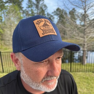 Op de afbeelding: Marineblauwe baseballcap met een gebogen klep. Een rechthoekige, bruine leren patch is op de voorkant genaaid, met de tekst "Reel Men Love Jesus". De pet is gemaakt van een stevige stof.