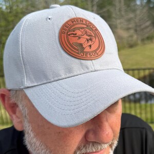 Op de afbeelding: Lichtgrijze baseballcap met een bruin leren embleem. Het embleem toont een visillustratie en de tekst "REEL MEN LOVE JESUS". De pet heeft een gebogen klep en een klassiek zes-panelen ontwerp.