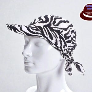 May include: A black and white zebra print visor cap. The cap is tied at the back and has a curved brim. The image also includes a logo with the text "ELITESHATU BRANSON UNIQUE DESIGNS HAT / CAPS".