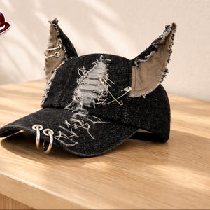 Puede incluir: Gorra de béisbol de mezclilla negra con detalles desgastados, dos aros plateados en la visera y detalles en forma de orejas. La gorra tiene un diseño rasgado con costuras visibles y alfileres de seguridad. El logo dice "UNIQUE DESIGNS HATS & CAPS".
