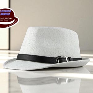 Può includere: Un cappello fedora bianco con fascia in pelle nera e fibbia argentata. Il cappello ha una tesa strutturata e una trama strutturata. Il logo in alto a sinistra dice "ELUVASHEET BRANSON UNIQUE DESIGNS HAT / CAPS".