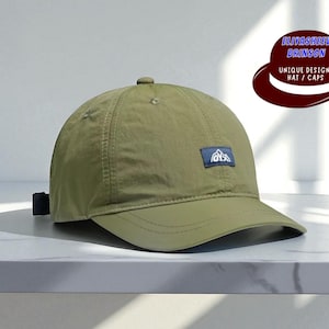 Może przedstawiać: Oliwkowozielona czapka z daszkiem z zakrzywionym daszkiem i małą kwadratową naszywką. Naszywka ma grafikę górską. Czapka ma czarny regulowany pasek. Tekst na zdjęciu brzmi "ELIYASHEEV BRINSON UNIQUE DESIGNS HAT / CAPS."