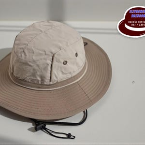 May include: A beige sun hat with a wide brim and a rounded crown. The hat features a drawstring and a contrasting band around the base of the crown. The hat is designed for sun protection and outdoor activities.