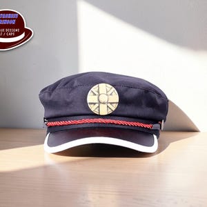 May include: Navy blue cap with a white brim and a gold emblem. A red rope detail accents the cap, which has the text "ELITESHOP CRIMSON UNIQUE DESIGNS HAT / CAPS" on a sticker in the upper left corner.