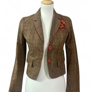 May include: Brown plaid blazer with a decorative patch on the upper left chest. The blazer has two front pockets and two buttons. The blazer is displayed on a mannequin.
