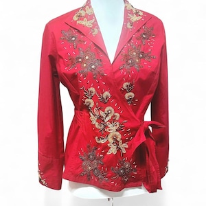 May include: A vibrant red wrap blouse with long sleeves and a tie closure. The blouse is adorned with intricate floral embroidery in gold, brown, and sequin accents, creating a decorative design. The collar and cuffs also feature embroidered details.