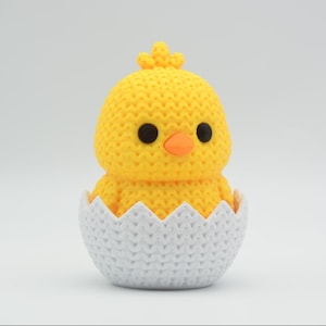 May include: A yellow knitted chick with black eyes and an orange beak emerging from a white knitted eggshell. The chick has a small crest on its head. The eggshell has a jagged edge.