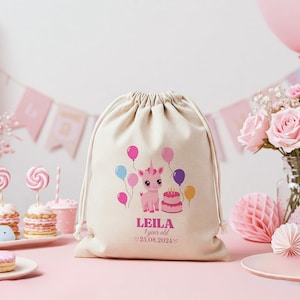 Może przedstawiać: Biała torba ze sznurkiem do ściągania z nadrukiem różowego jednorożca i balonów. Torba ma nadrukowane imię "LEILA" i tekst "1 year old 25.08.2024".