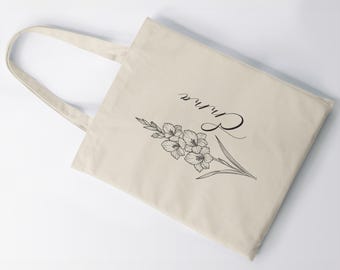 Puede incluir: Bolso tote color crema con un diseño floral negro y la palabra "Rhina" en escritura cursiva. El diseño presenta flores de gladiolo y hojas, ofreciendo un aspecto elegante y artístico. El bolso tiene asas largas.