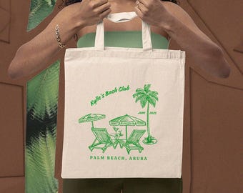 May include: Cream-coloured tote bag featuring a green design of beach chairs, parasols, and a palm tree. The text on the bag says "Kylie's Bach Club" and "Palm Beach, Aruba, June 2025". It has two white handles.