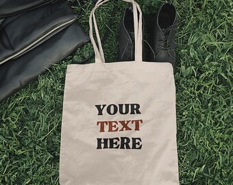 Puede incluir: Una bolsa de lona blanca con asas negras. La bolsa tiene el texto "YOUR TEXT HERE" impreso en la parte delantera en letras negras y marrones.