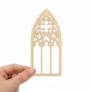 May include: A laser-cut wooden gothic window design. The window features a pointed arch and intricate floral tracery. The natural wood color provides a neutral base for crafting or decoration. The window has three vertical panels.