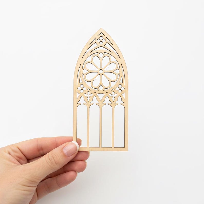May include: A laser-cut wooden decorative piece in the shape of a cathedral window. The window features intricate floral and geometric patterns, with a pointed arch and vertical panels. The natural wood color provides a neutral tone.