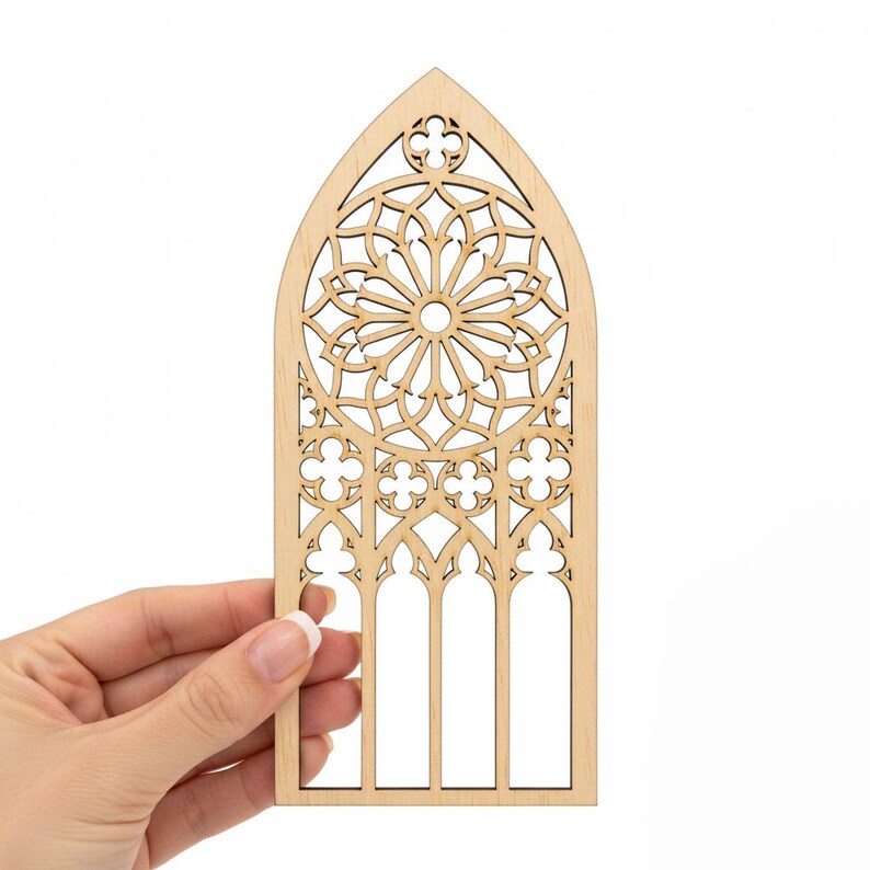 May include: A laser-cut wooden decorative piece in the shape of a cathedral window. The window features intricate geometric patterns and a central circular design. The wood has a natural, light brown color, and the piece is held by a hand against a white background.